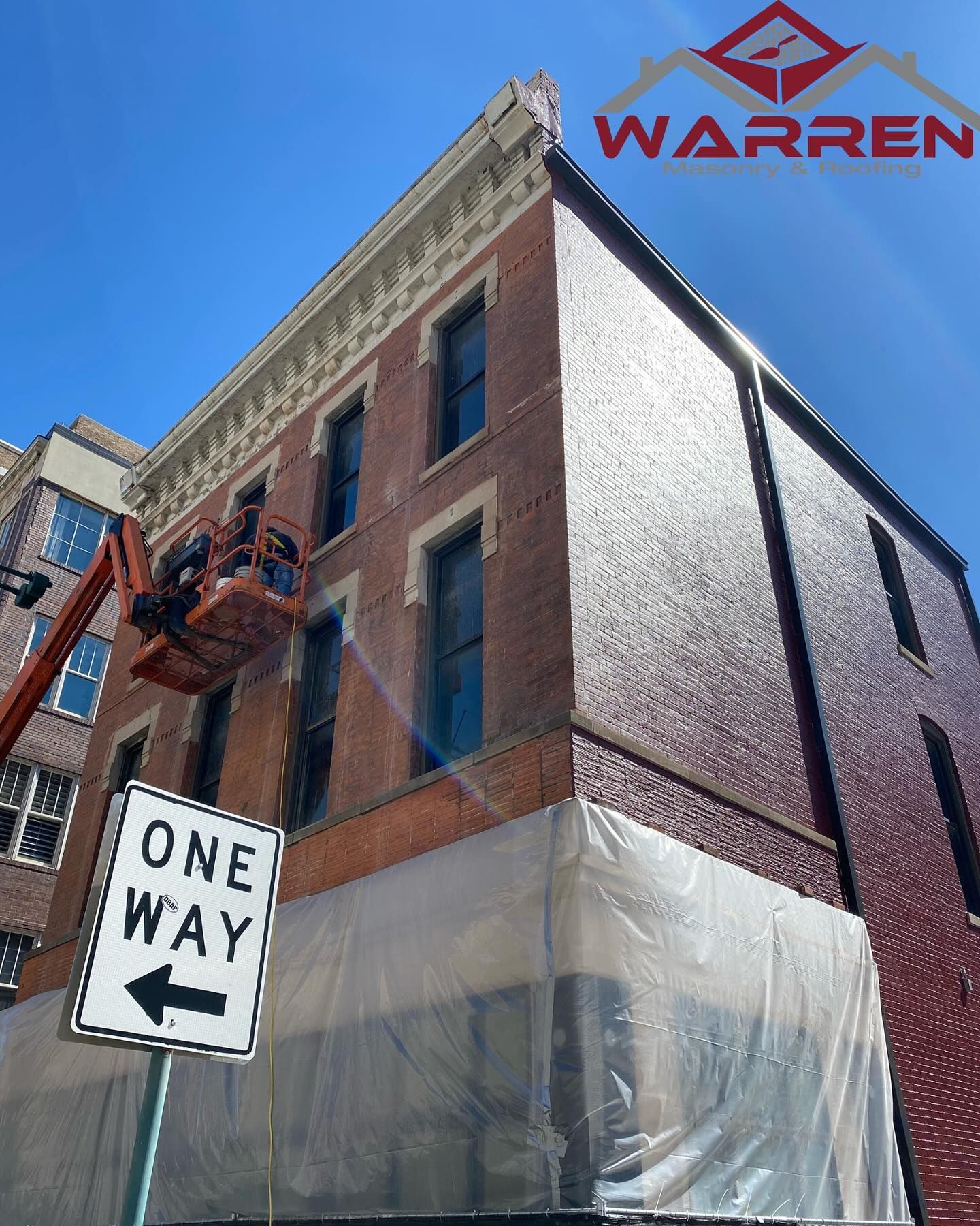 A person in a lift works on the facade of a historic brick building while a one-way street sign stands in the foreground.