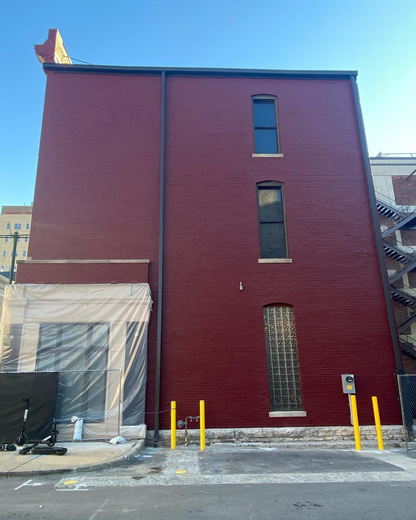 A three-story dark red brick building with three vertical windows, a ground-level plastic barrier, and yellow bollards.