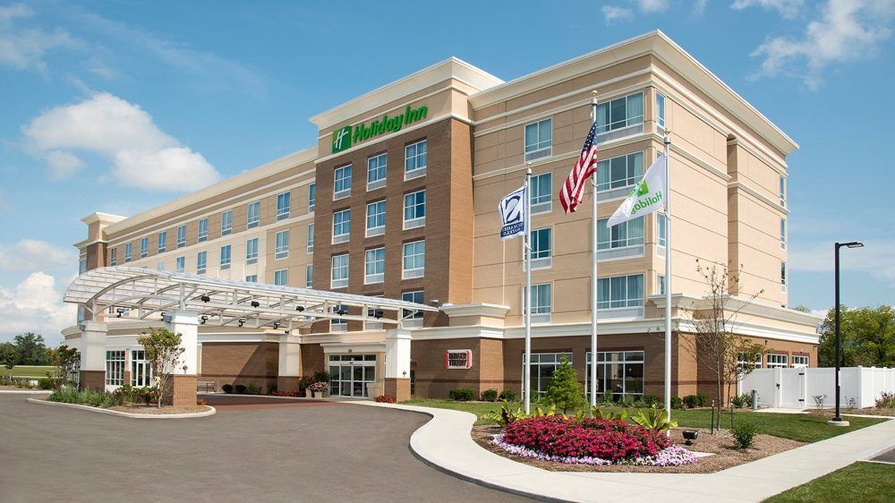 A Holiday Inn hotel exterior with a paved entrance, flags, and a small garden area on a sunny day with a blue sky.