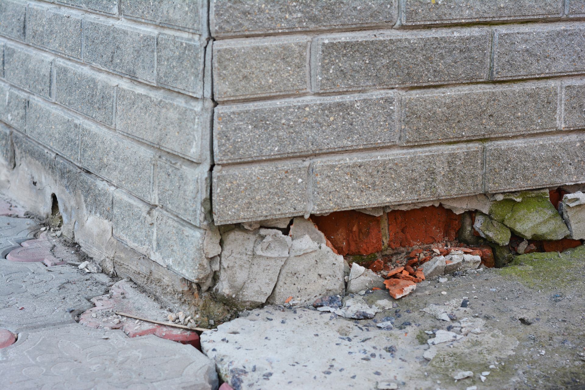 A corner of a building exterior showing crumbled mortar and damaged masonry at the base where it meets the ground.
