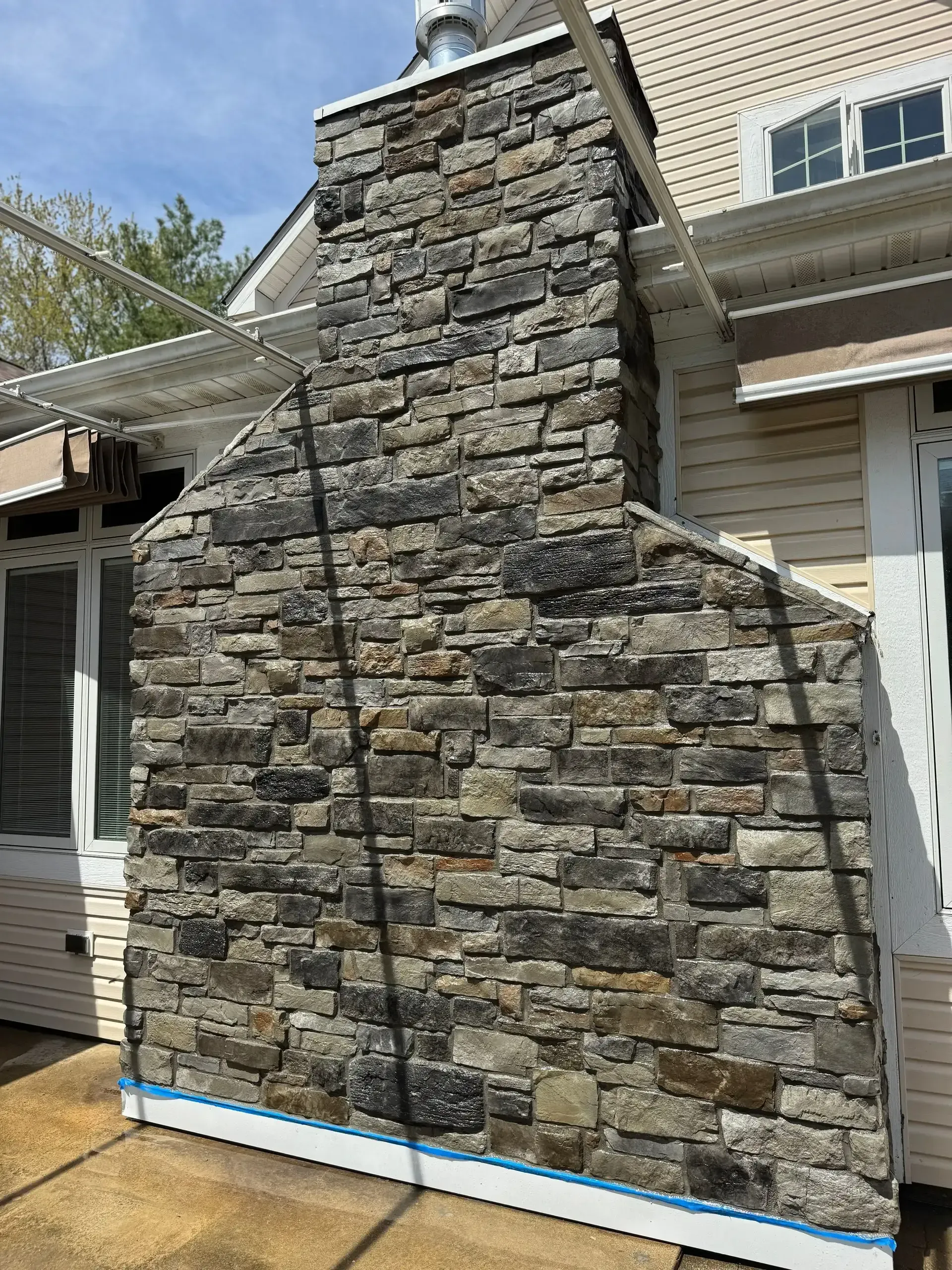 Stone-clad chimney or exterior wall section under construction beside a house, with blue foam at the base.