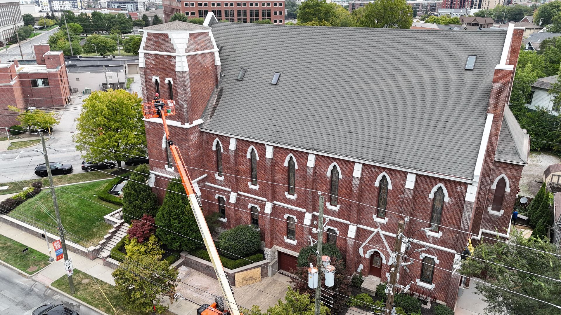 An aerial view of a red brick church with a tall steeple, being serviced by a construction lift on a sunny day.