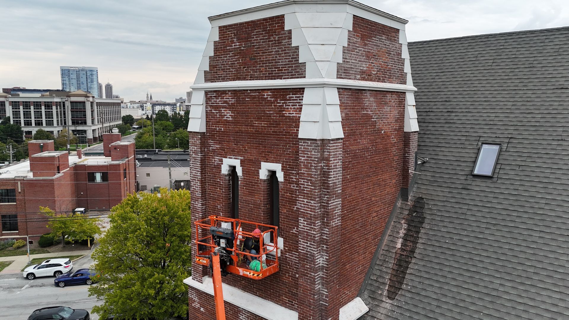 An aerial view shows workers in an orange lift platform repairing the brick tower of a church near a city skyline.