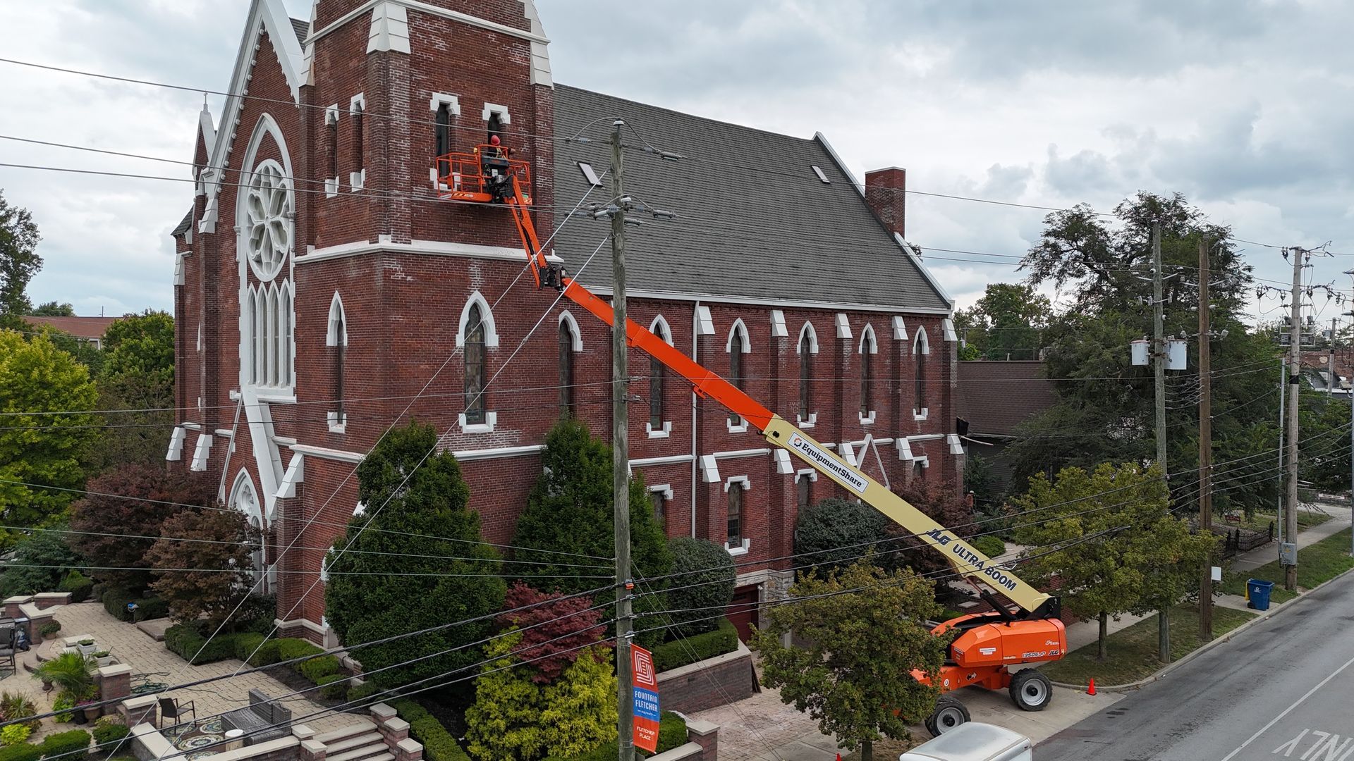 Workers in an orange boom lift reach a brick tower on a building rooftop, with city buildings visible in the background.