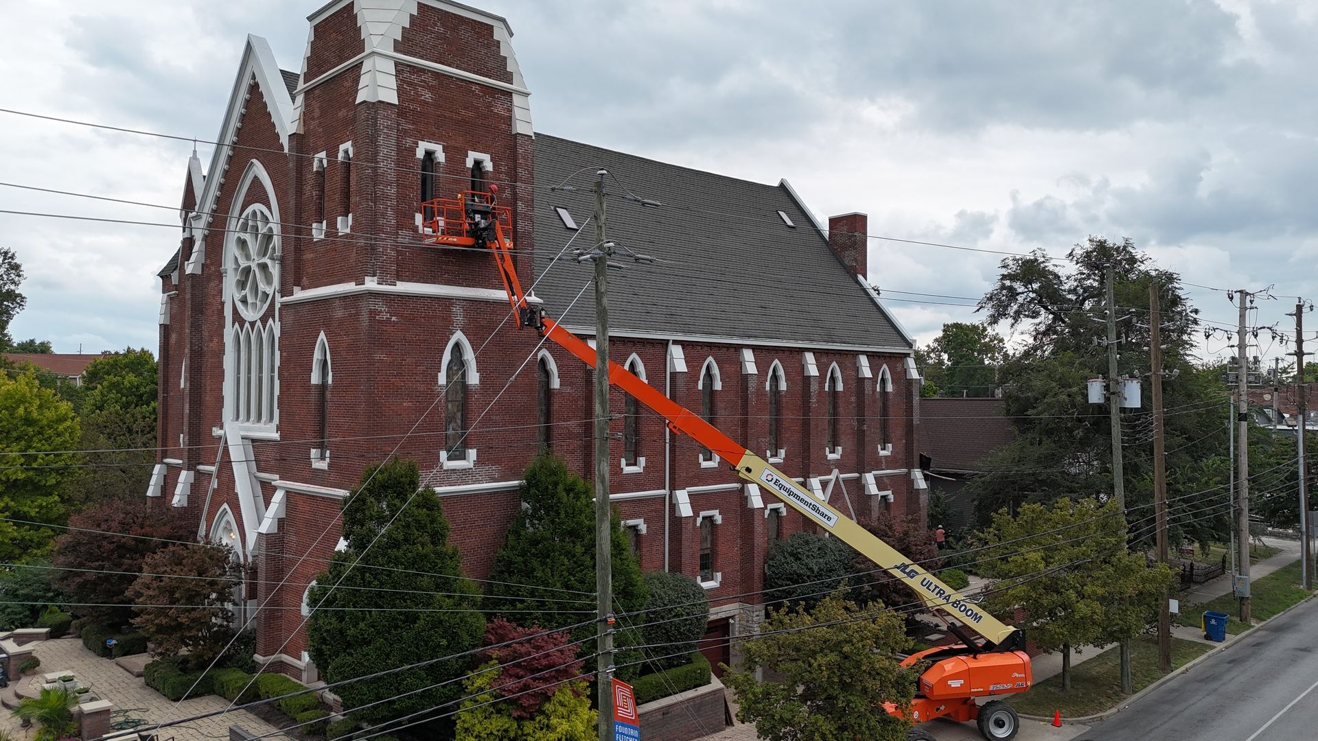 A worker in a boom lift reaches the upper windows of a historic red brick church near a road and utility poles.