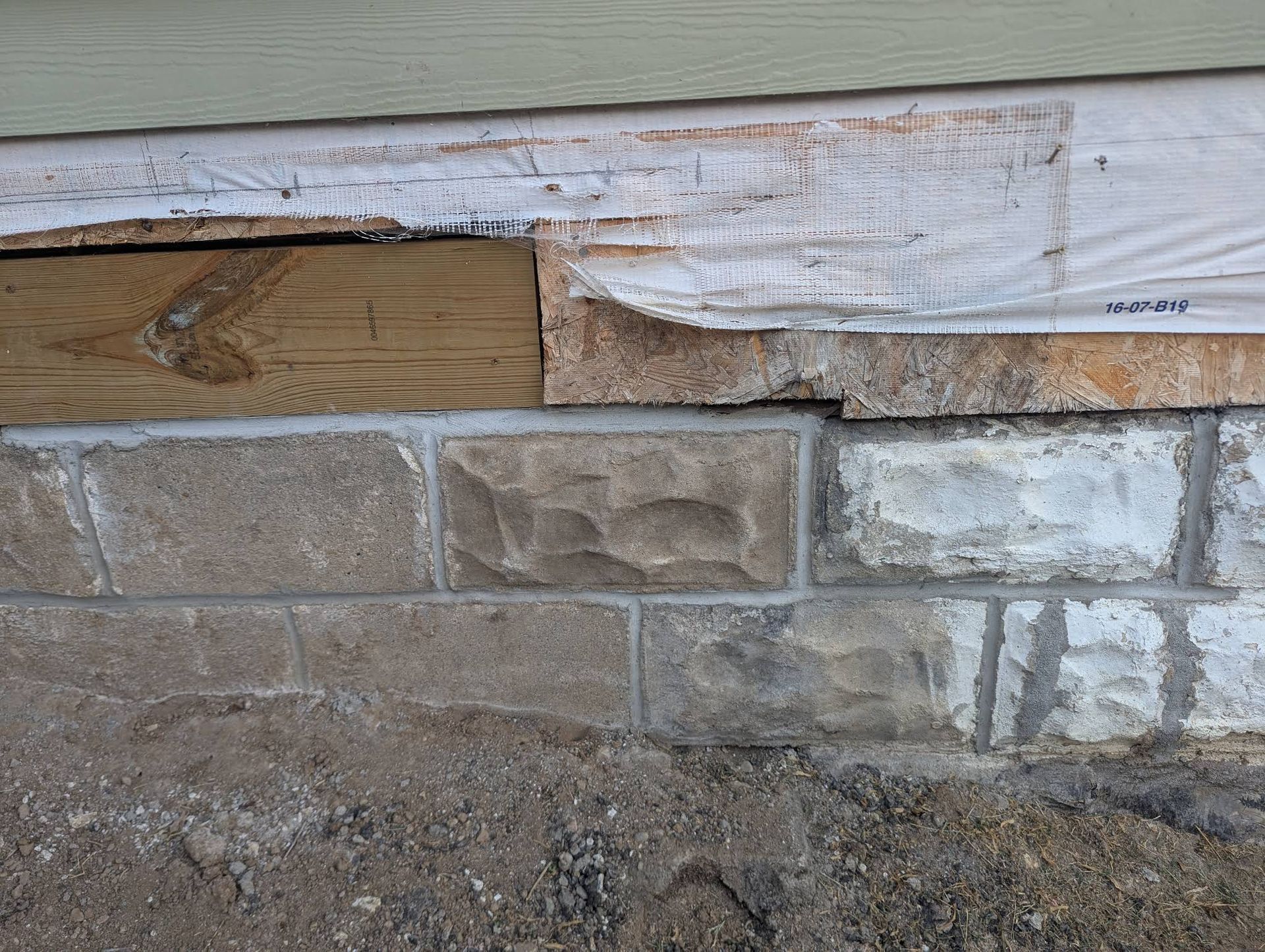 A close-up view of an exterior wall foundation showing damaged wooden boards, torn house wrap, and concrete masonry units.